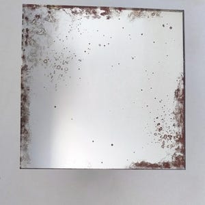 Könnte beinhalten: Quadratischer Spiegel mit einem antiken, verwitterten Aussehen. Die versilberte Oberfläche hat dunkelbraune, marmorierte Ränder und verstreute dunkle Flecken, die eine Vintage-Ästhetik erzeugen. Der Rahmen ist nicht sichtbar. Der Spiegel ist etwa 30 cm groß.