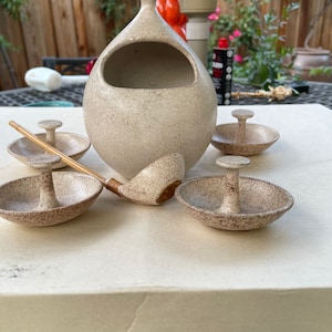 May include: A set of five ceramic ring holders with a matching ceramic pitcher. The ring holders are beige with brown speckles and have a small, raised knob in the center. The pitcher is beige with brown speckles and has a large opening at the top. The set is arranged on a white surface.