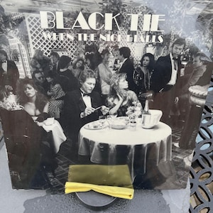 May include: Black and white album cover titled "Black Tie When the Night Falls" featuring a group of people at a formal event. A yellow cloth and a black disc are in the foreground.