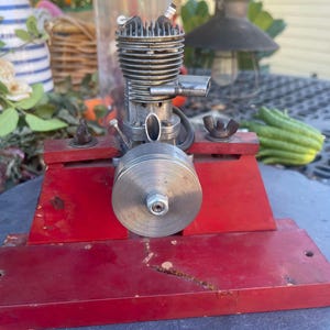 May include: A metallic model engine with a silver-colored cylinder head and a circular flywheel, mounted on a rectangular red base. The engine features a small spark plug at the top and is secured to the base with two metal wing nuts.