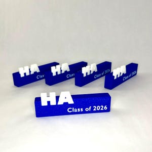 May include: A collection of blue and white graduation favors. Each rectangular block features the letters "HA" in white, with "Class of 2026" printed in white. The blocks are arranged on a white surface.