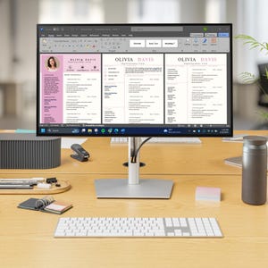 May include: A computer monitor displays a resume template with the name "Olivia Davis" in a pink and white design. The monitor sits on a wooden desk with a keyboard, mouse, and other office supplies. A small plant and a gray speaker are also on the desk.