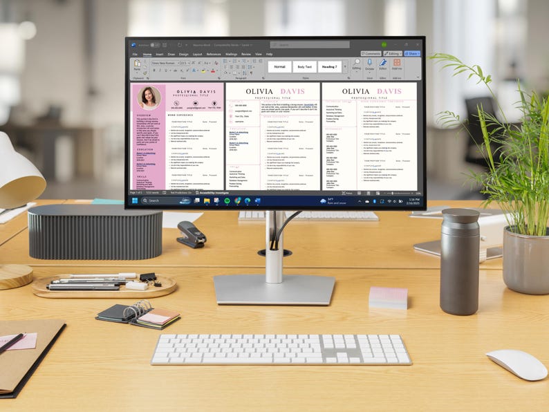 May include: A computer monitor displays a resume template with the name "Olivia Davis" in a pink and white design. The monitor sits on a wooden desk with a keyboard, mouse, and other office supplies. A small plant and a gray speaker are also on the desk.