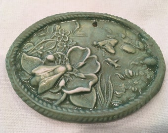 May include: A green ceramic oval ornament with a raised design of flowers and bees. The ornament has a hole at the top for hanging.