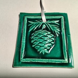 Può includere: Piastrella quadrata in ceramica verde smeraldo con un design a rilievo di pigna e ramo. La piastrella ha una finitura lucida e un nastro bianco per appenderla. La pigna è centrata all'interno di un bordo.