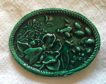 May include: A green ceramic oval plaque with a raised design of flowers, leaves, and bees. The plaque has a rope-like border.
