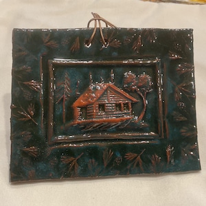 May include: A rectangular ceramic wall plaque with a glossy dark teal glaze featuring a relief-carved orange cabin in a forest setting. The border is decorated with embossed pine needles and cones, and a leather cord is attached for hanging.
