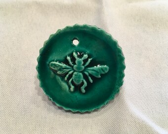 May include: A teal green ceramic ornament with a raised bee design. The ornament has a hole at the top for hanging.