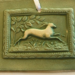 May include: A square, green ceramic ornament with a raised relief design. The design features a running dog within a rectangular frame, surrounded by foliage. A white ribbon is attached for hanging.