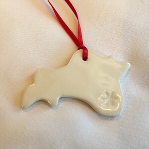 Puede incluir: Adorno de cerámica blanca con forma de perro con una huella de pata en el lateral. El adorno tiene una cinta roja atada en la parte superior.