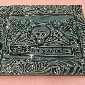 May include: A teal blue ceramic tile with a raised design of a skull with wings, crossed bones, and the text "MEMENTO MORI".