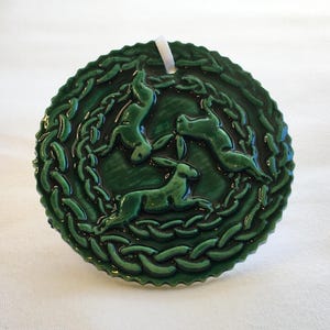 May include: A round, dark green ceramic ornament featuring three hares in a circular design. The hares are arranged in a triangle, with a braided border around them. A white ribbon is attached for hanging.