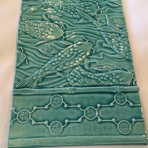 Pode incluir: Azulejo de cerâmica azul-turquesa com um design em relevo de peixes koi nadando na água. O azulejo tem uma borda de bambu e um padrão repetitivo de círculos estilizados.