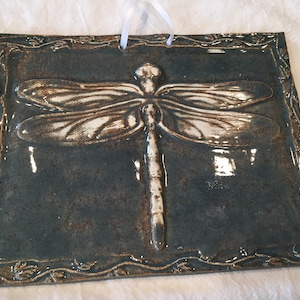 Puede incluir: Un colgante de pared de cerámica con un diseño de libélula blanca sobre un fondo azul verdoso. La libélula está en relieve y tiene un diseño detallado. El fondo tiene una textura moteada y un esmalte marrón. El colgante de pared tiene una cinta blanca simple para colgar.