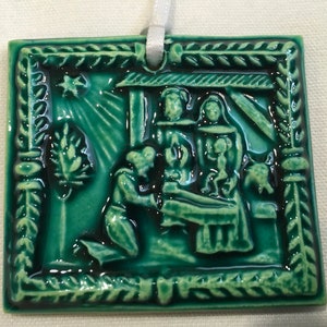 May include: A green ceramic Christmas ornament depicting the nativity scene. The ornament features a star, a manger, and figures of Mary, Joseph, and baby Jesus.