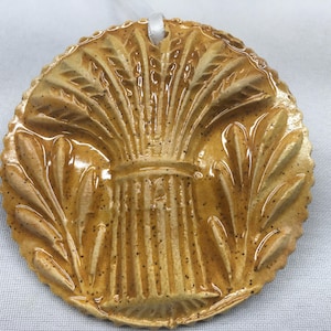 May include: A round, ceramic ornament with a raised design of wheat stalks in a bundle. The ornament is glazed in a light brown color with a speckled finish.