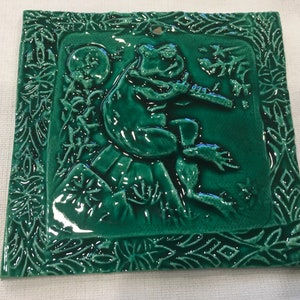 May include: A green ceramic tile with a raised design of a frog playing a flute. The tile is surrounded by a decorative border of leaves and flowers.