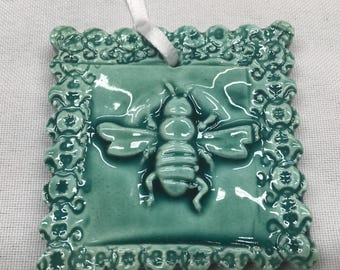 May include: A teal blue ceramic ornament with a raised bee design. The ornament has a white string for hanging and a decorative border.