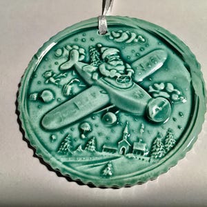 May include: A teal ceramic Christmas ornament with a scalloped edge. The ornament features a relief design of Santa Claus flying a biplane over a winter landscape with trees, a church, and clouds. A white ribbon is attached for hanging.