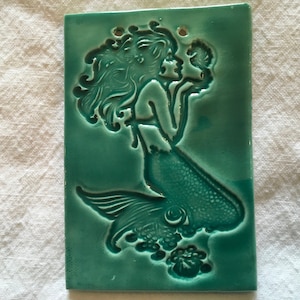 Peut inclure: Carreau de céramique vert d'eau avec un motif de sirène. La sirène a des cheveux flottants, un coquillage et une queue détaillée. La tuile a deux trous en haut pour l'accrocher.