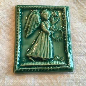 May include: A green ceramic tile featuring a raised image of an angel holding a wreath of flowers. The angel is wearing a long dress and has wings.