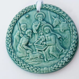 Puede incluir: Un adorno de cerámica azul verdoso que representa la escena de la natividad. El adorno presenta una estrella, un pesebre, un bebé Jesús, María, José, un pastor y dos animales.