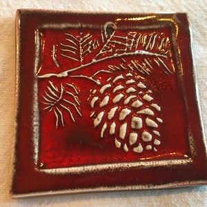 May include: A red ceramic tile with a white raised design of a pine cone and branch.