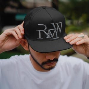 May include: Black baseball cap with a white embroidered "RWM" logo. The cap is being held by a person wearing a white t-shirt. The hat has a structured crown and a flat brim.