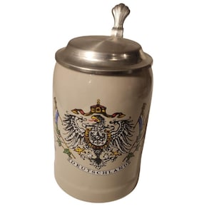 May include: A beige ceramic beer stein featuring a metal lid with a shell-shaped thumb lift. The front displays a colorful eagle crest with the word DEUTSCHLAND printed in black capital letters on a banner beneath the central emblem.