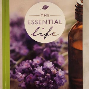 May include: A book titled "The Essential Life" with a lavender and brown color scheme. The cover features a circular design with the title and a sprig of lavender. A brown glass bottle is visible on the right side of the book.