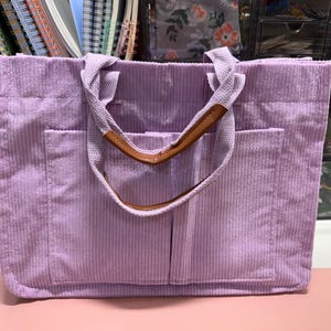 May include: A lavender corduroy tote bag with two front pockets and sturdy handles. The handles are a light canvas material with a brown leather accent. The bag is designed for carrying items and has a rectangular shape.