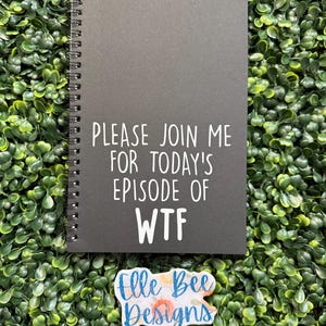 May include: A black spiral-bound notebook with white text that reads "PLEASE JOIN ME FOR TODAY'S EPISODE OF WTF." A colorful sticker with the text "Elle Bee Designs" is below the notebook. The background is a dense arrangement of green leaves.