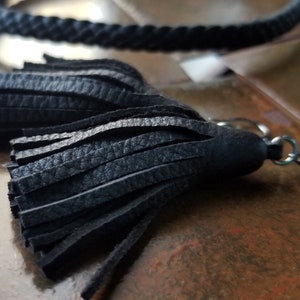 Puede incluir: Primer plano de un llavero con borla de cuero negro con un cierre plateado. La borla está hecha de múltiples tiras de cuero texturizado. En el fondo se ve un cordón negro trenzado.