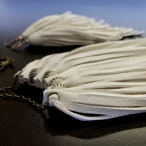 May include: A pair of white leather tassel earrings with a silver chain. The tassels are long and flowing, and the chain is attached to a small silver hoop.