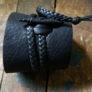 May include: A black leather cuff bracelet with a braided leather accent. The cuff is thick and wraps around the wrist. A braided leather cord and knot detail the top of the cuff.