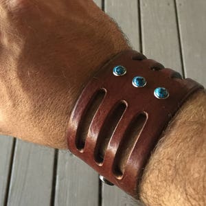 May include: A brown leather cuff bracelet with turquoise stones. The bracelet has three cutouts and is worn on a person's wrist.