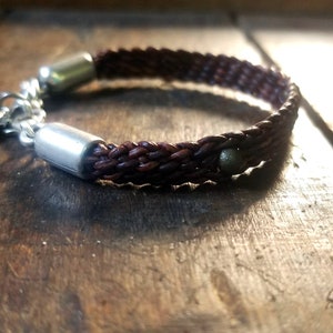 Puede incluir: Un brazalete de cuero marr&oacute;n con un dije de pluma de plata y dos acentos de metal plateado. El brazalete est&aacute; trenzado y tiene una cuenta marr&oacute;n oscura.