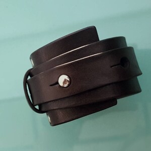 Puede incluir: Un brazalete de cuero negro con un tachuela de plata. El brazalete está hecho de varias capas de cuero y tiene un diseño simple y minimalista.