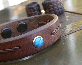 Puede incluir: Un brazalete de cuero marrón con un diseño trenzado y un acento de piedra turquesa. El brazalete tiene un cierre magnético.