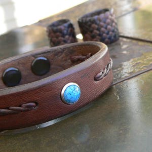 May include: A brown leather bracelet with a braided design and a turquoise stone accent. The bracelet has a magnetic clasp.