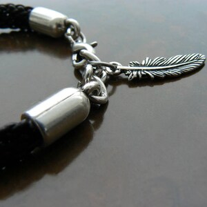 Puede incluir: Un brazalete de cuero trenzado negro con una cadena de plata y un dije de pluma de plata.