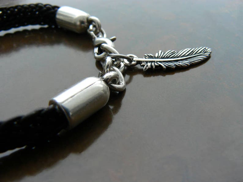 Puede incluir: Un brazalete de cuero trenzado negro con una cadena de plata y un dije de pluma de plata.
