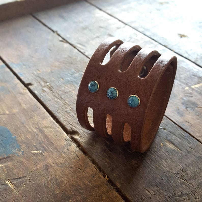 May include: Brown leather cuff bracelet with three turquoise stones. The bracelet has a cut-out design.