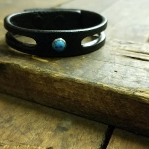 Puede incluir: Un brazalete de cuero negro con un acento de piedra turquesa. El brazalete tiene un dise&ntilde;o de doble bucle.