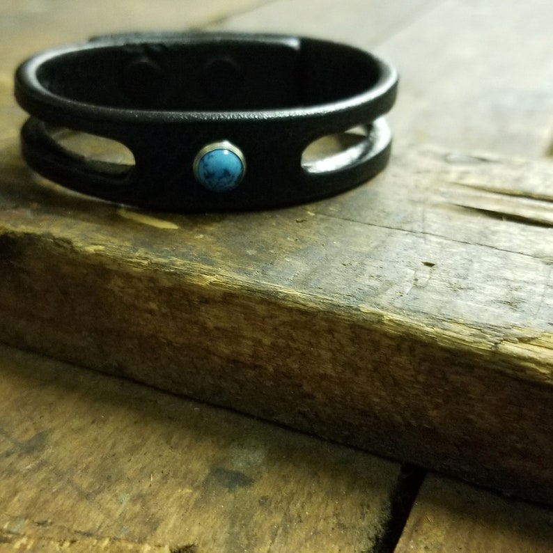 Puede incluir: Un brazalete de cuero negro con un acento de piedra turquesa. El brazalete tiene un dise&ntilde;o de doble bucle.