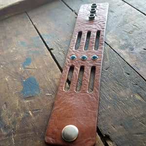May include: A brown leather bracelet with turquoise stones and silver accents. The bracelet has a cut-out design and is adjustable.