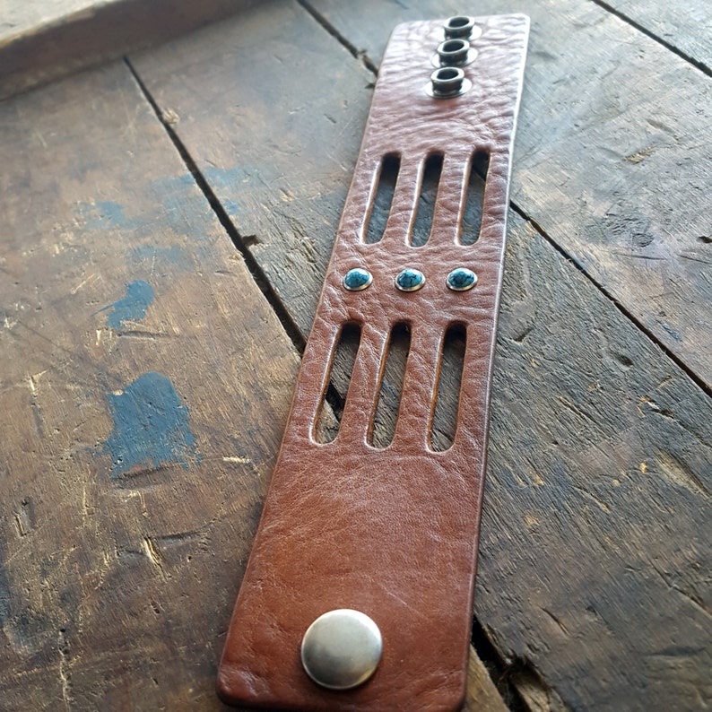 May include: A brown leather bracelet with turquoise stones and silver accents. The bracelet has a cut-out design and is adjustable.