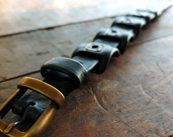May include: Close-up of a black leather bracelet with a brass buckle. The bracelet is lying on a weathered wooden surface. The buckle is a warm gold color, and the leather appears to be slightly worn.