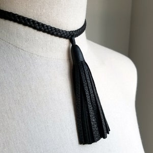 Puede incluir: Un collar gargantilla de cuero negro con un borla. La borla está hecha del mismo cuero que la gargantilla y cuelga del centro de la gargantilla.
