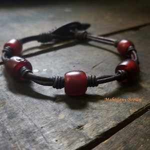 May include: A leather bracelet with red glass beads and a brown leather closure. The bracelet is on a wooden surface. "Mahogany Brown" is written on the surface.